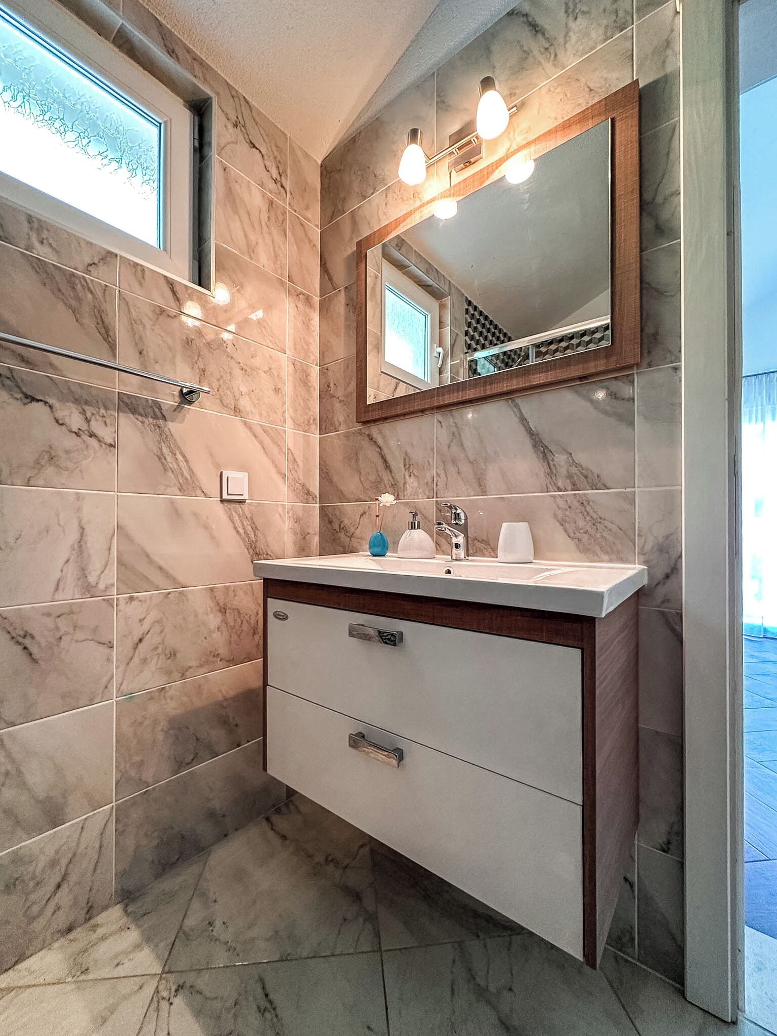 A bathroom with marble tiles, a vanity with a mirror, and a window.