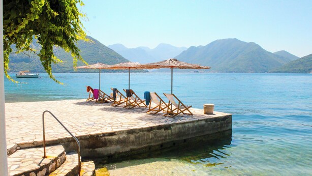 A dock with lounge chairs and umbrellas, a boat in the background, and mountains in the distance. - Apartments Stella del Mare (Risan)