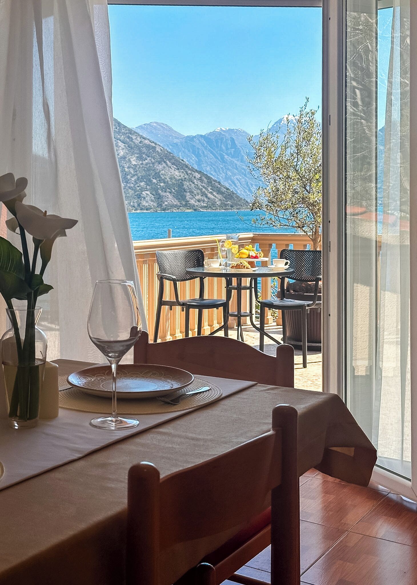 A dining area with a table set for two, a vase with flowers, and a view of the sea and mountains.