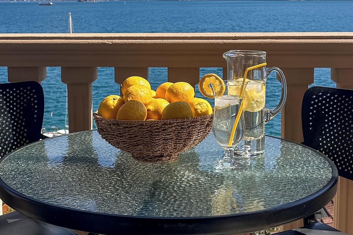 Une table avec une carafe en verre et un panier de citrons, donnant sur un lac avec des montagnes en arrière-plan.