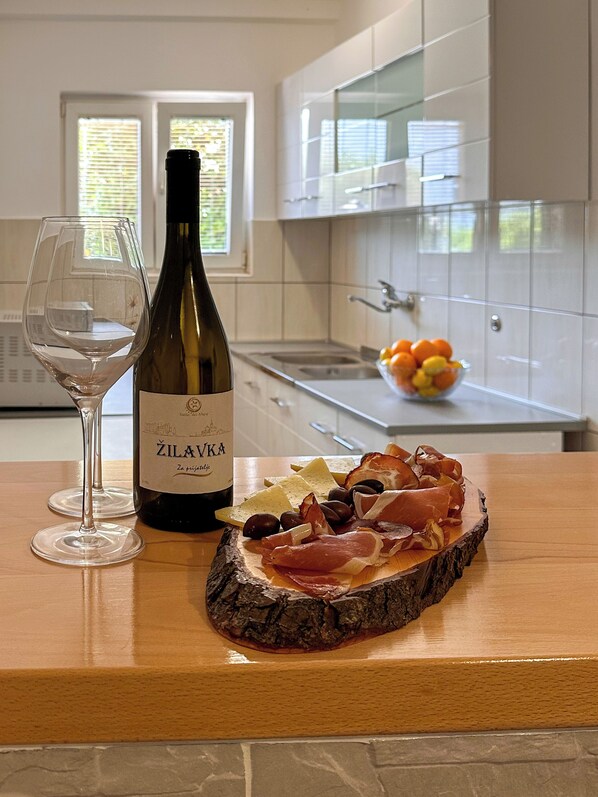 Una botella de vino Zilavka, una copa y una tabla de fiambres con queso, aceitunas y embutidos sobre una tabla de madera en una cocina con armarios blancos y una ventana.