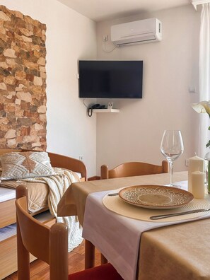 A room with a stone wall, a bed, a TV mounted on the wall, a dining table with a candle, and a vase with flowers. - Apartments Stella del Mare (Risan)