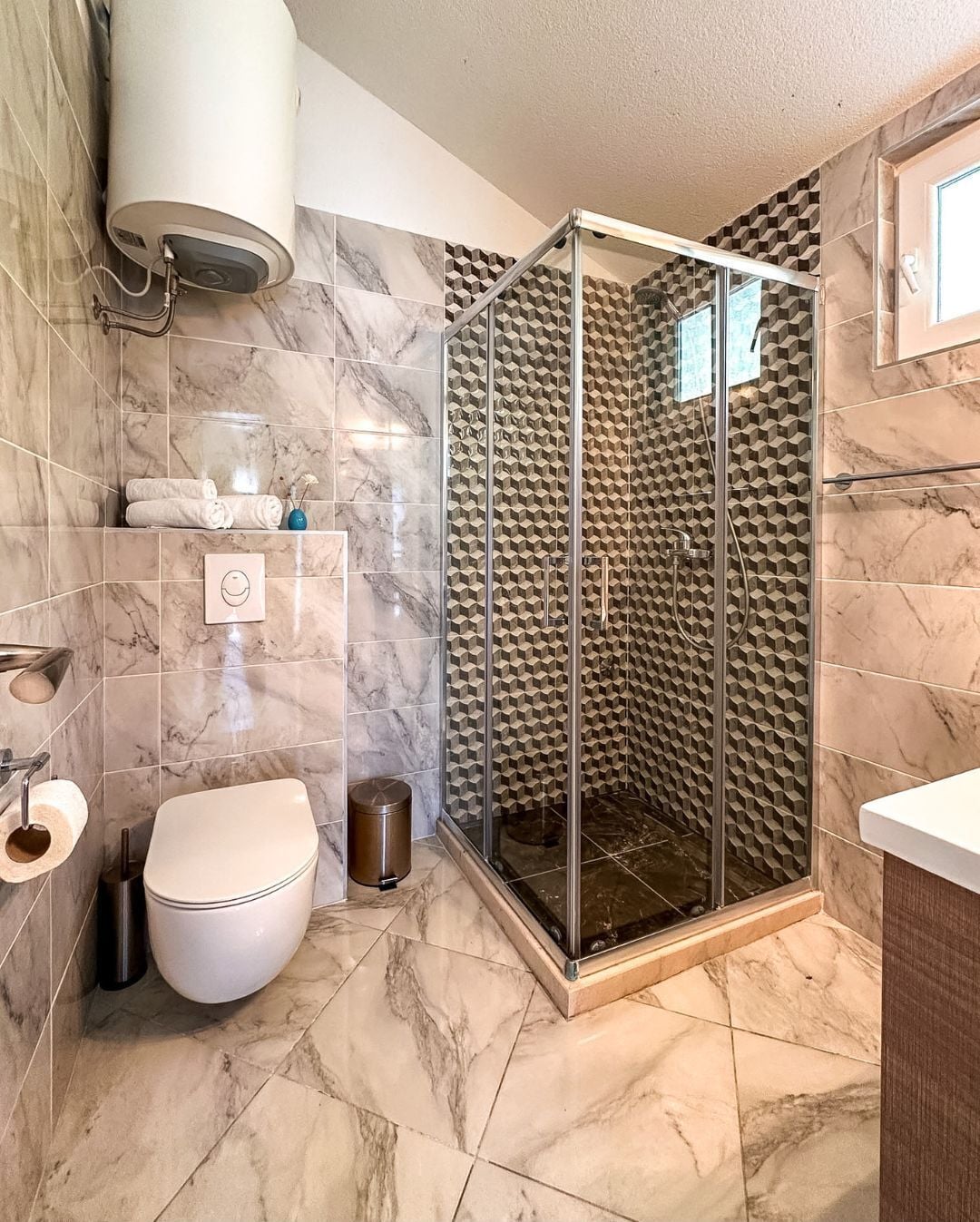 A bathroom with a glass-enclosed shower, marble tiles, a toilet, and a towel rack.