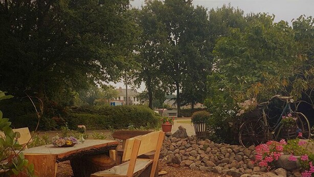 Un área de estar al aire libre de estilo rústico con una mesa y sillas de madera, rodeada por un muro de piedra y vegetación.