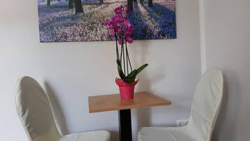 Ein Zimmer mit zwei weißen Stühlen, einem kleinen Tisch mit einer Orchidee im Topf und einem Waldgemälde an der Wand.