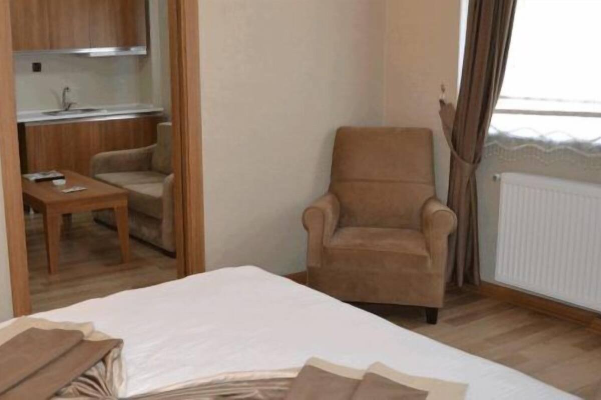 Ein Schlafzimmer mit einem Bett, einem Sessel, einem kleinen Tisch und einer Küchenzeile im Hintergrund.