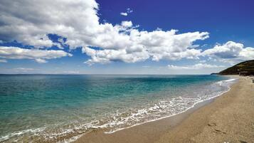Ein Strand mit klarem türkisfarbenem Wasser, einem sandigen Ufer und einer entfernten hügeligen Landschaft unter einem teils bewölkten Himmel.