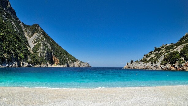 A beach with clear turquoise water, rocky cliffs, and a pebbly shore. - Corali Hotel (Kymi-Aliveri)