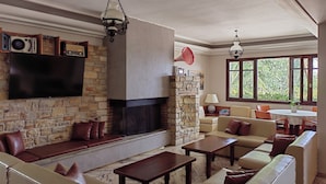 A living room with a stone fireplace, leather sofas, a flat-screen TV, and a dining area with a table and chairs. - Corali Hotel (Kymi-Aliveri)