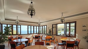 A spacious dining area with multiple round tables and chairs, large windows offering a view of the sea, and hanging chandeliers. - Corali Hotel (Kymi-Aliveri)