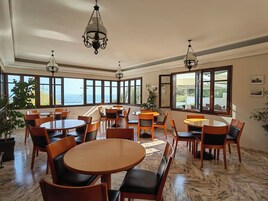 Un amplio comedor con varias mesas redondas y sillas, ventanas grandes con vistas al mar y candelabros colgantes.