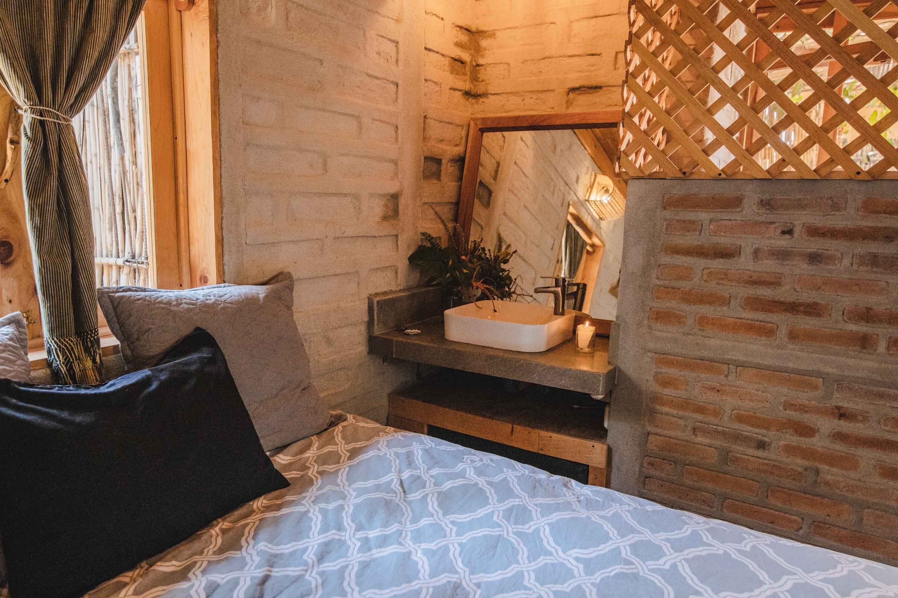 A cozy bedroom with a brick wall, a wooden bed frame, a bedside table with a mirror, and a window with curtains.