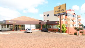 Un hôtel avec une enseigne indiquant « nossa casa » et « HOTEL CHURRASCARIA ». Deux voitures sont garées devant, et le bâtiment a un toit plat et une façade en briques.