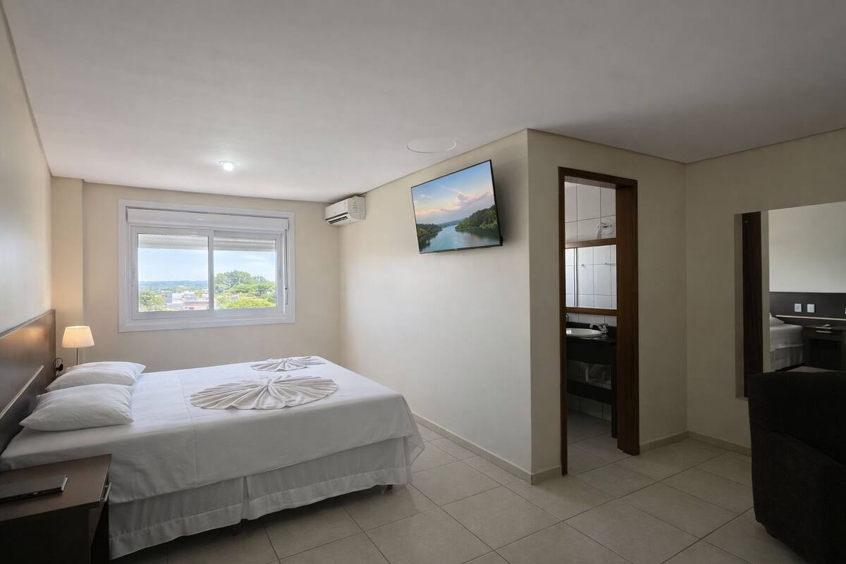 Un dormitorio con cama, televisor, ventana con vistas y baño visible a través de una puerta abierta.