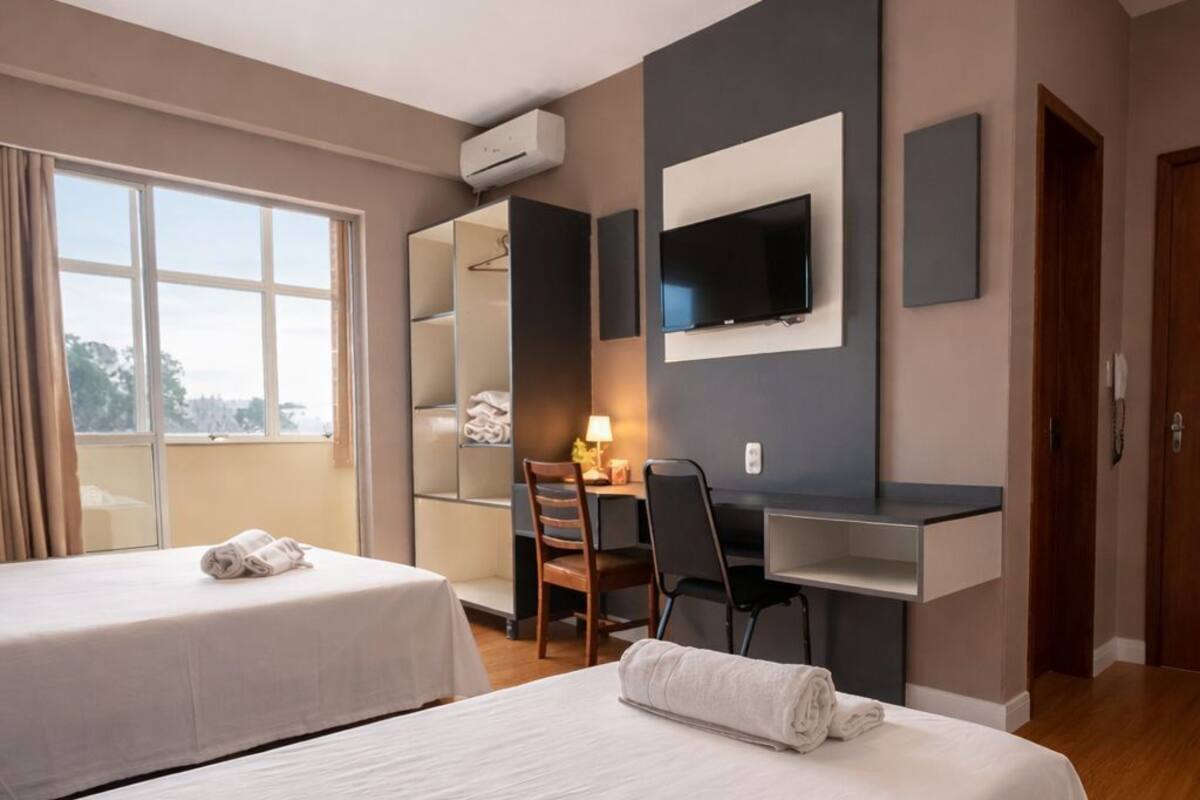 Habitación de hotel con dos camas, un escritorio con silla, un televisor montado en la pared y un ventanal con cortinas.