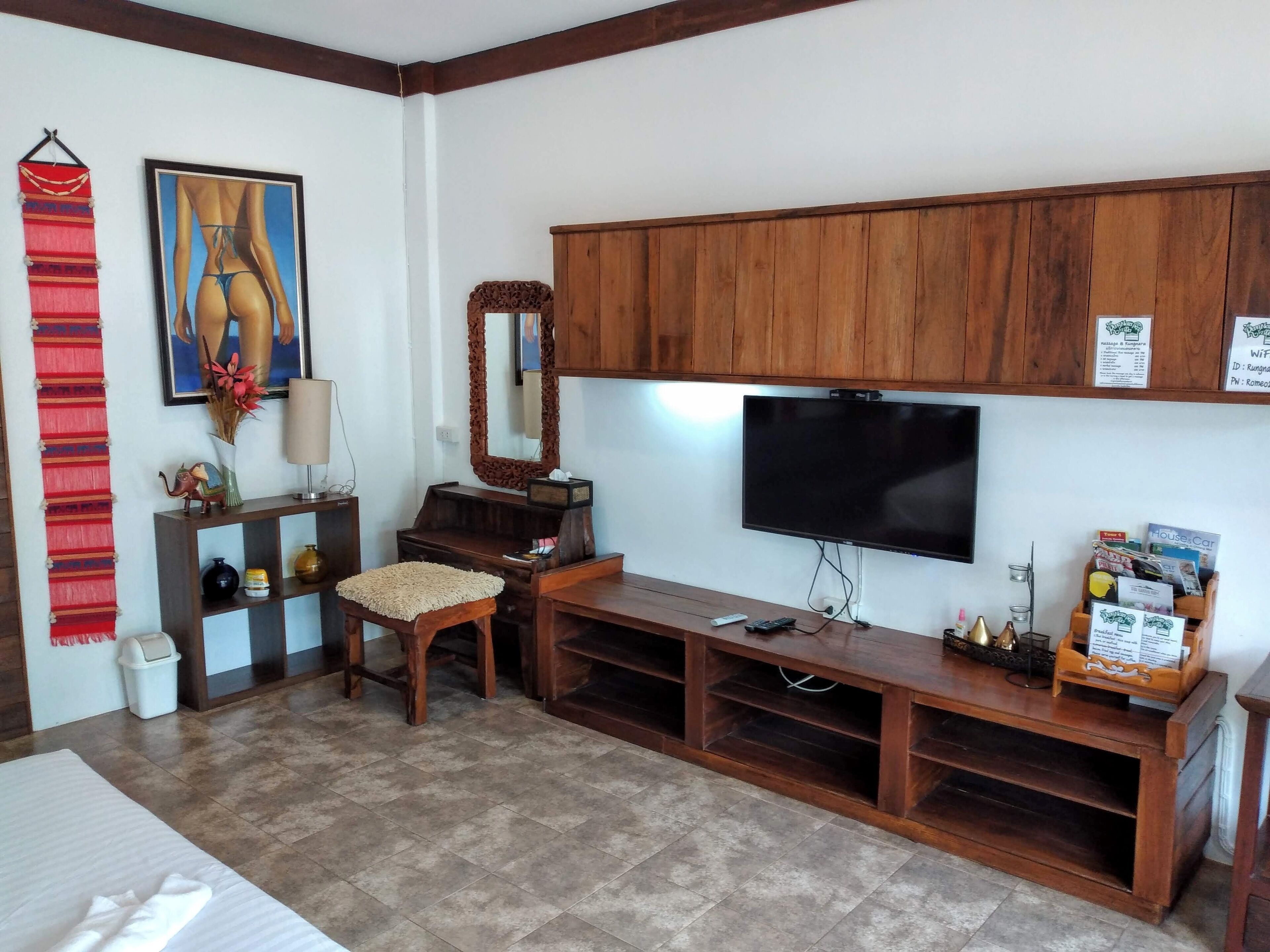 A living room with a TV, wooden cabinets, a bench, a small table with a lamp, and a red decorative wall hanging.