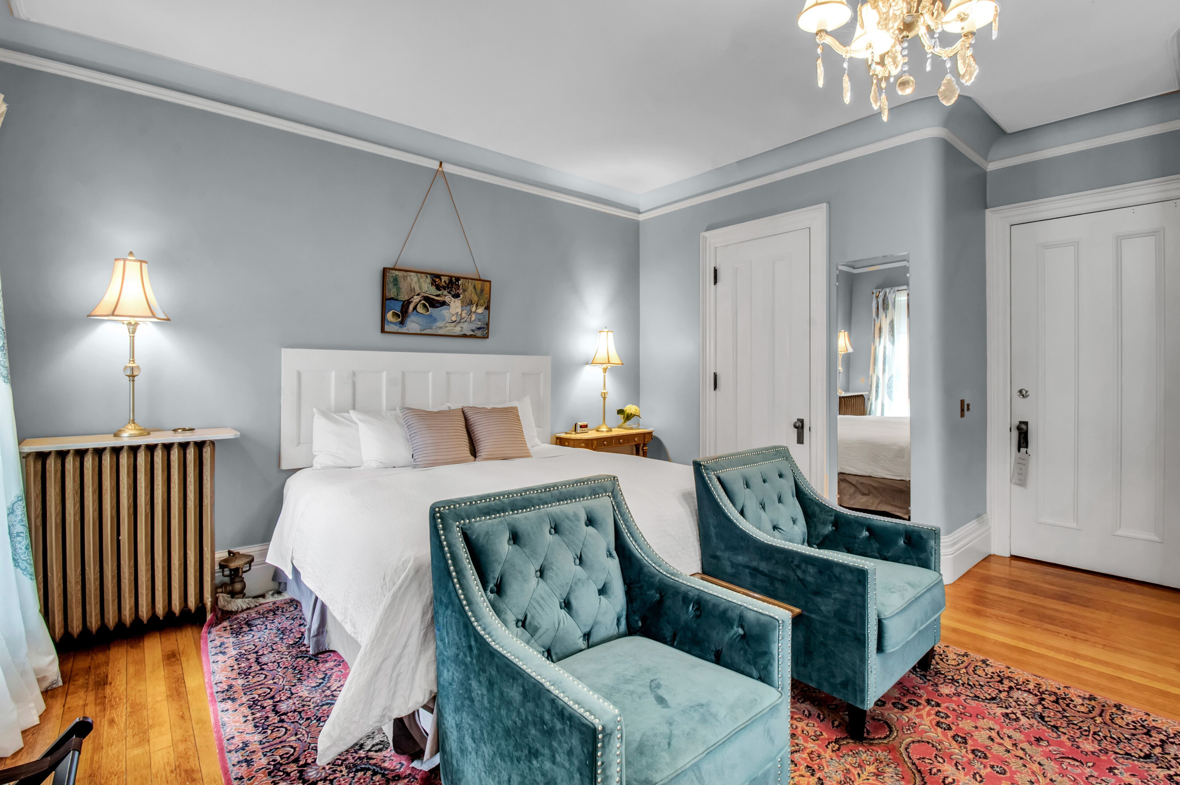 A bedroom with a large bed, two upholstered chairs, a radiator, and a chandelier.