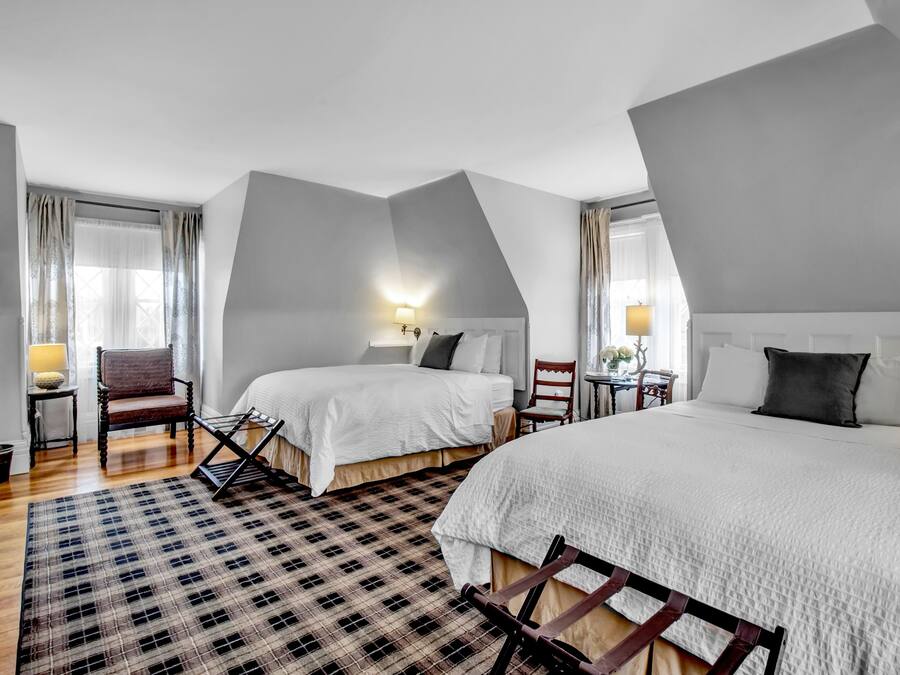 Ein Hotelzimmer mit zwei Betten, einem Schachbrettteppich, einem Holzstuhl, einem kleinen Tisch mit Lampe und einem Fenster mit Vorhängen.