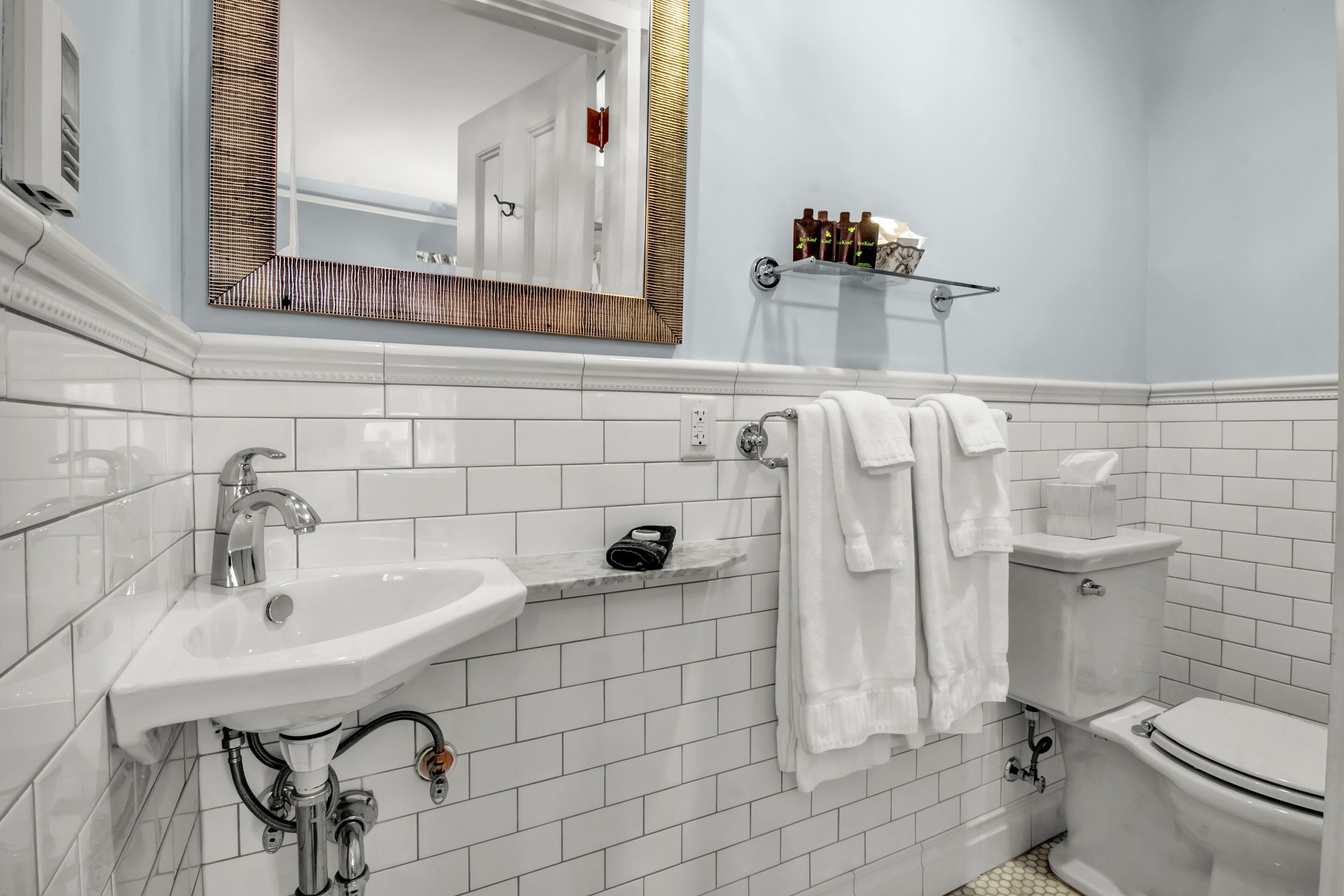 A modern bathroom with white tiles, a sink, a mirror, a toilet, and a shelf with toiletries.