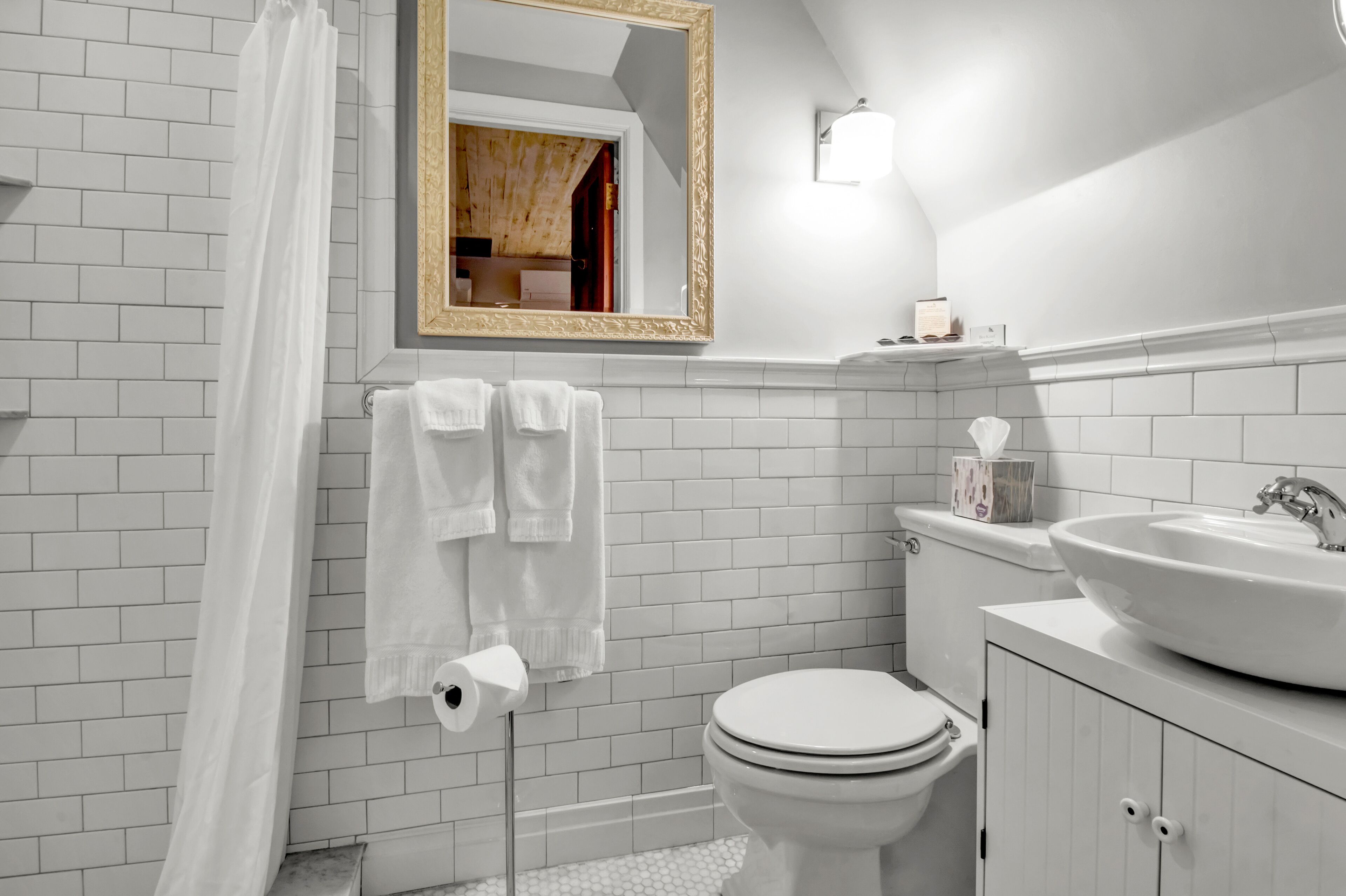 Un baño con paredes de azulejos blancos, un lavabo blanco, un inodoro y una cortina de ducha.