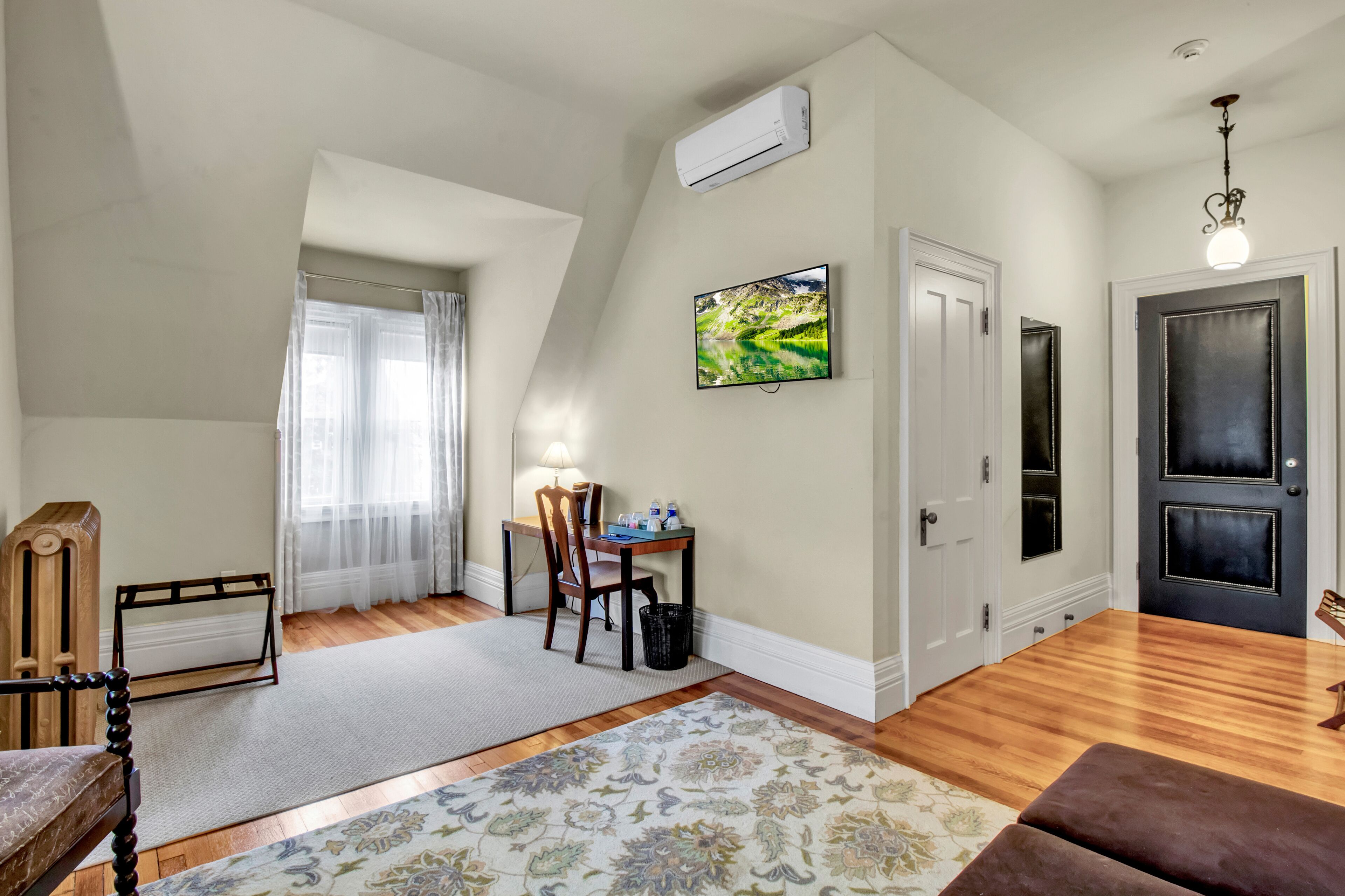 A room with a wooden floor, a desk with a chair, a lamp, a TV on the wall, and a door.