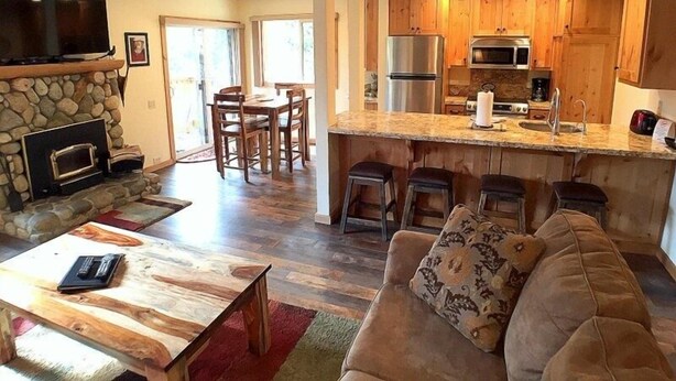 A spacious living room with a stone fireplace, a TV, and a wooden coffee table. Adjacent is a kitchen with wooden cabinets, a stainless steel refrigerator, and a bar area with stools. - 113 Standard Condo (Mammoth Lakes)