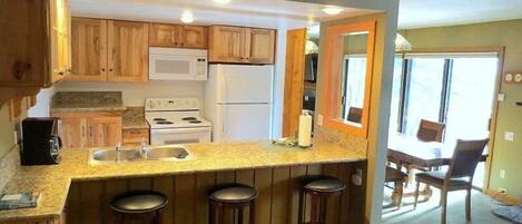 A compact kitchen with wooden cabinets, a microwave, a stove, and a refrigerator. There are three stools at the counter and a dining area with chairs and a table.