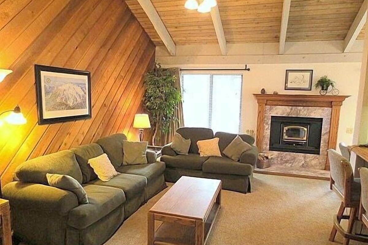 Ein gemütliches Wohnzimmer mit Kamin, zwei grünen Sofas, einem Holztisch und einer großen Pflanze.