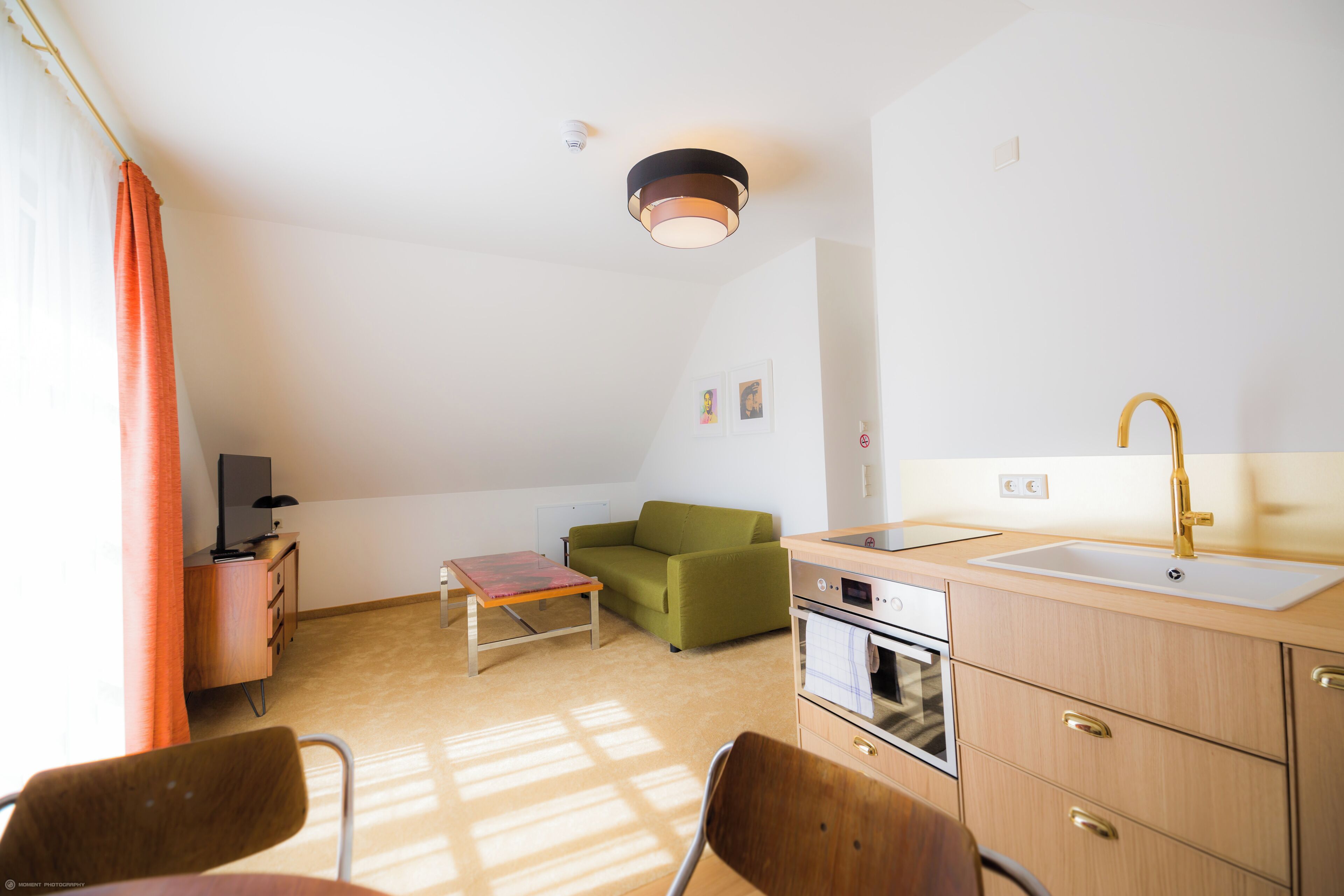 A compact room with a kitchenette, a green sofa, a wooden table, and a TV.