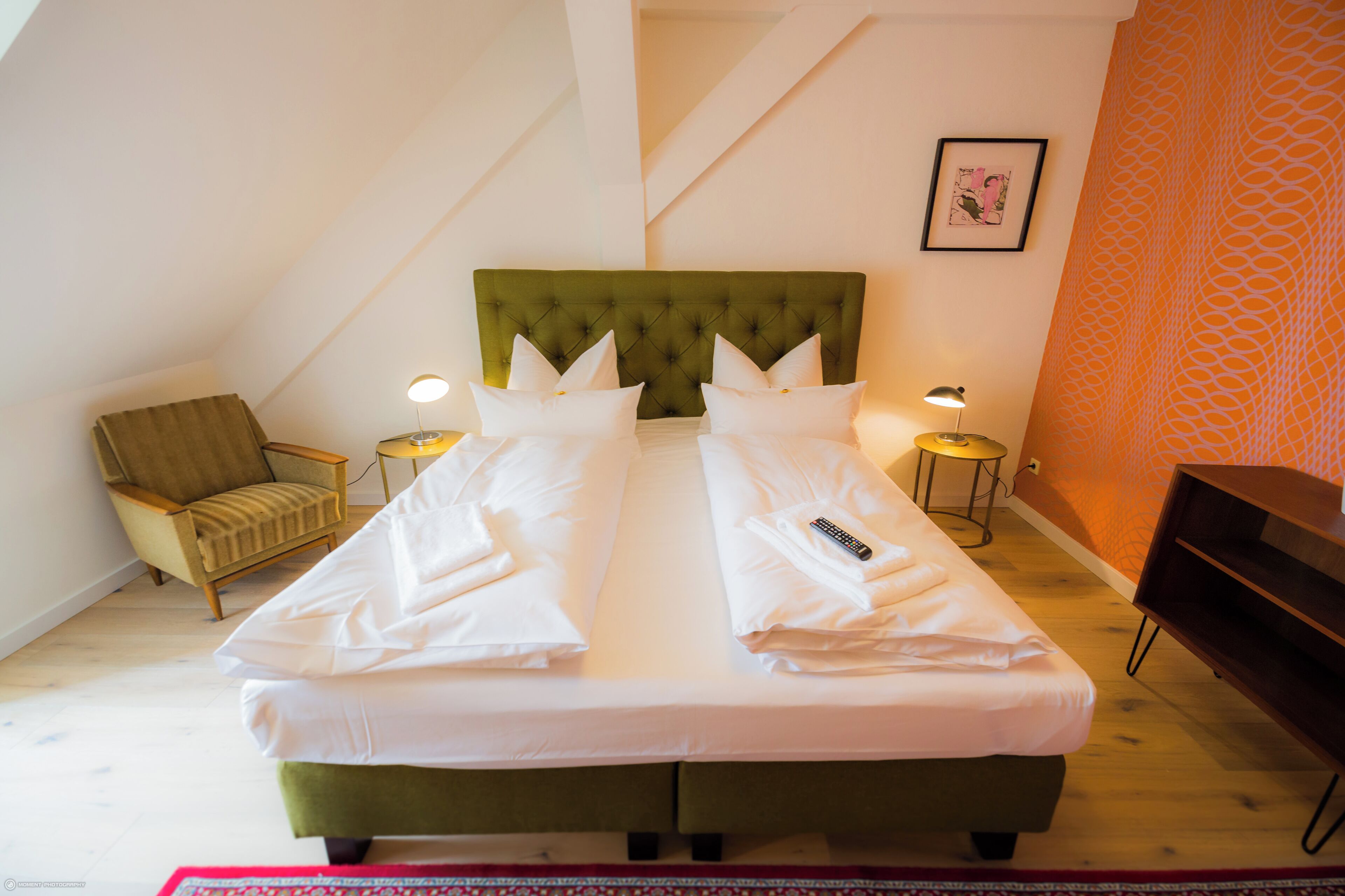 A double bed with a green upholstered headboard, two bedside lamps, a chair, and a framed picture on the wall.