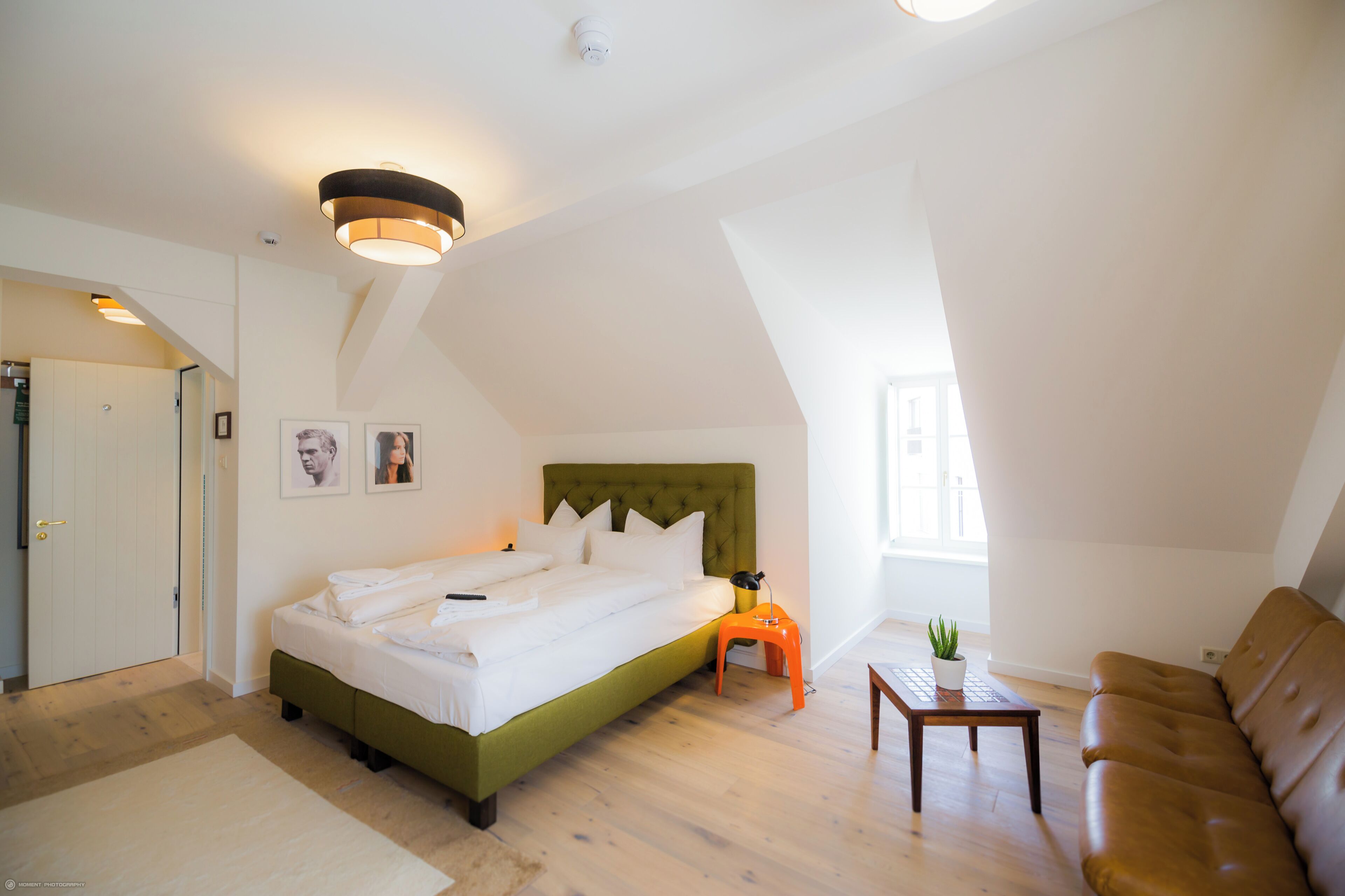 A bedroom with a bed, a green headboard, a small table with a plant, a brown leather sofa, and a door leading to another room.