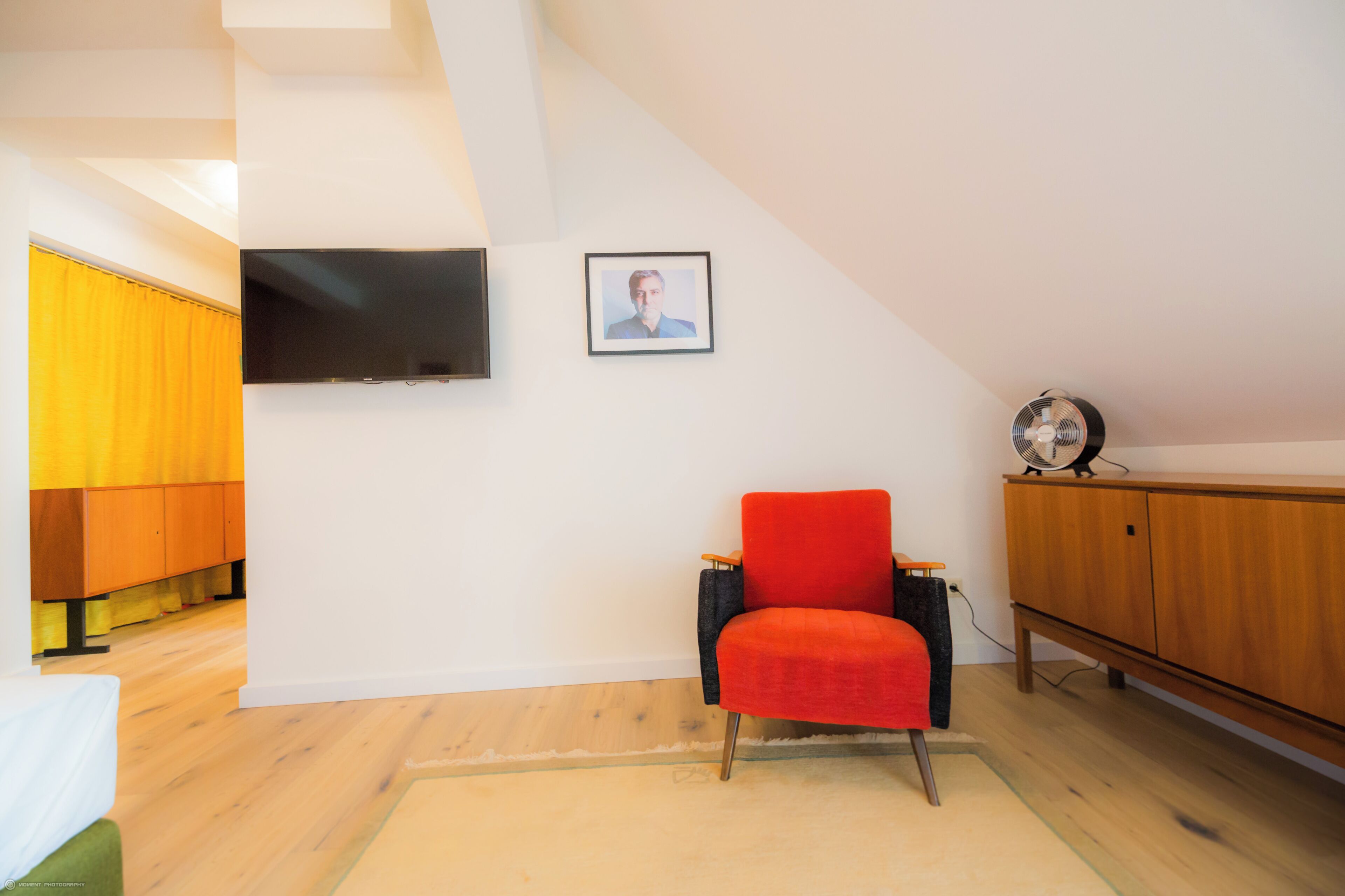 Una habitación con un televisor montado en la pared, una silla roja, un armario de madera y una cortina amarilla.