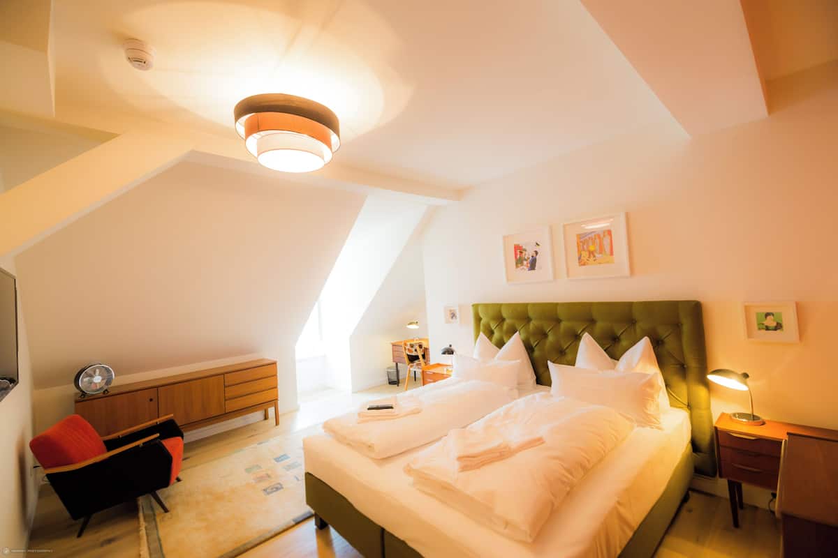Un dormitorio con cama, escritorio, silla, lámpara y obras de arte en las paredes.