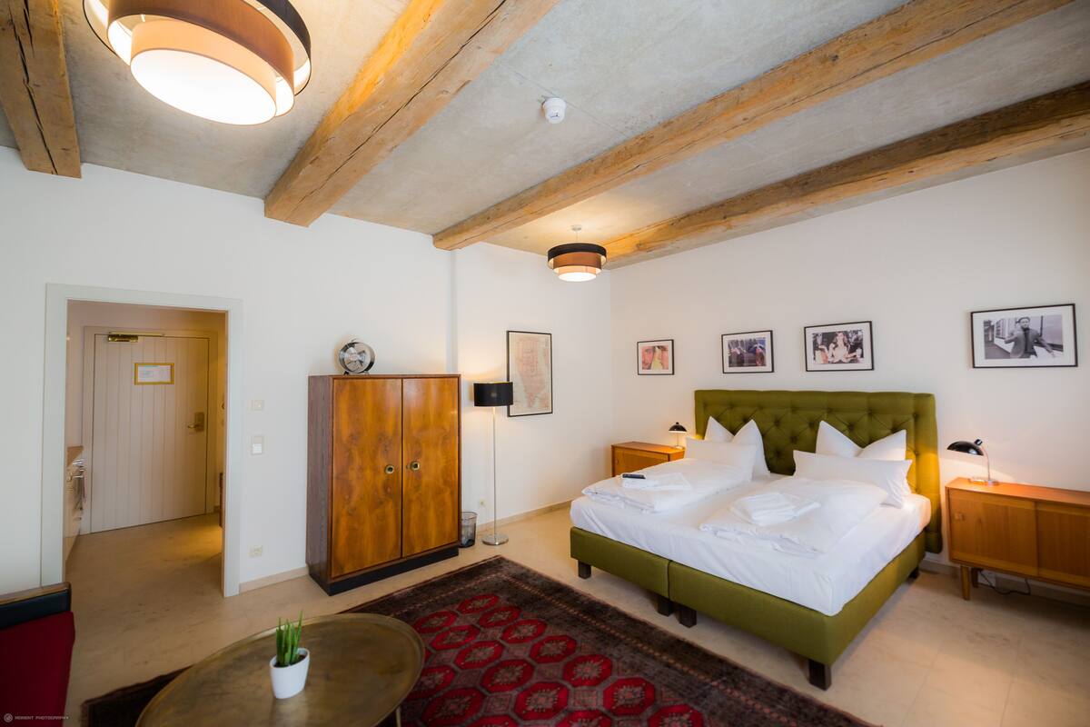 Un dormitorio con techo de madera, una cama tapizada de verde, un tapete rojo estampado, un armario de madera y una pequeña mesa con una planta.