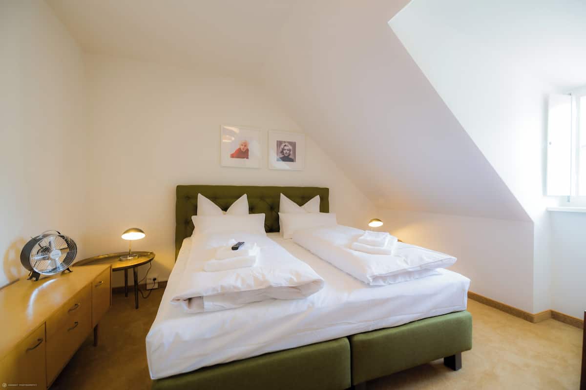 Una cama bien hecha con almohadas y un cabecero verde en una habitación con una mesita y un ventilador.