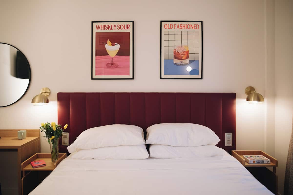 Una camera d'albergo con un letto grande, due quadri appesi al muro, un comodino con una lampada e un tavolino con un libro e un vaso di fiori.