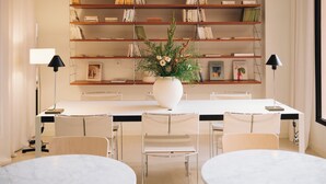 A modern dining area with a white table, four chairs, a white vase with flowers, and a bookshelf with books and decorative items. - H La Paloma Boutique (Barcelona)