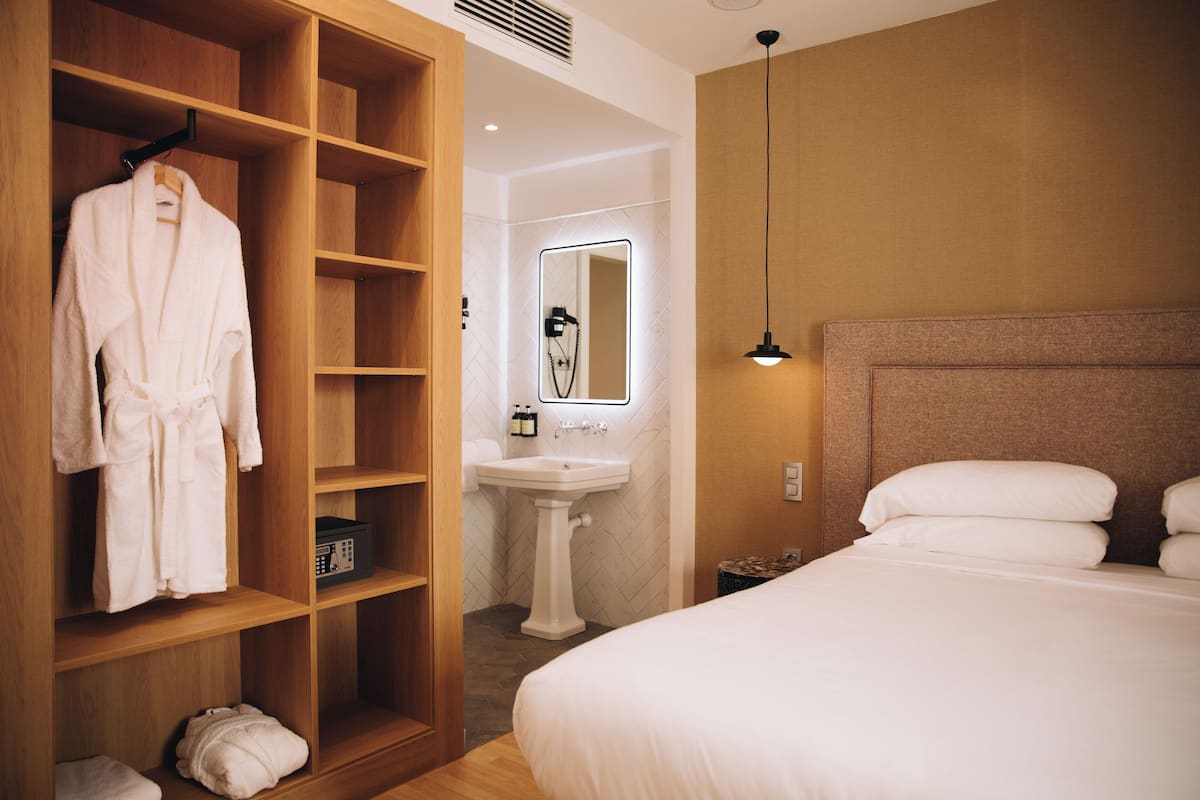 Una camera d'albergo con un armadio in legno, un letto bianco, un comodino e un bagno con lavandino e specchio.