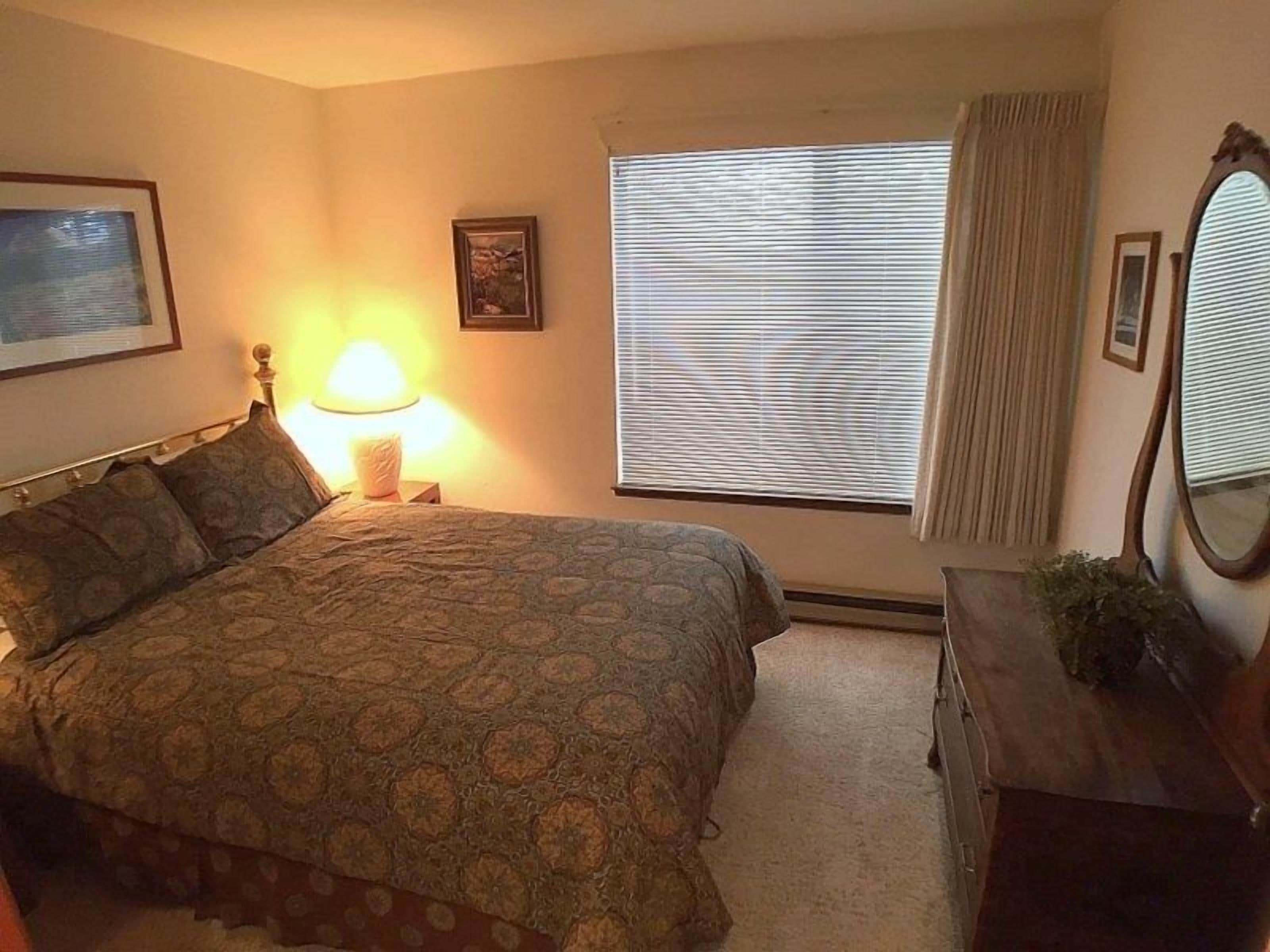 Un dormitorio con cama, lámpara de noche, ventana con persianas, cómoda y un cuadro en la pared.