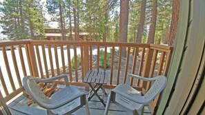 A wooden deck with two plastic chairs and a table, surrounded by tall trees. - 141 Premium Condo (Mammoth Lakes)