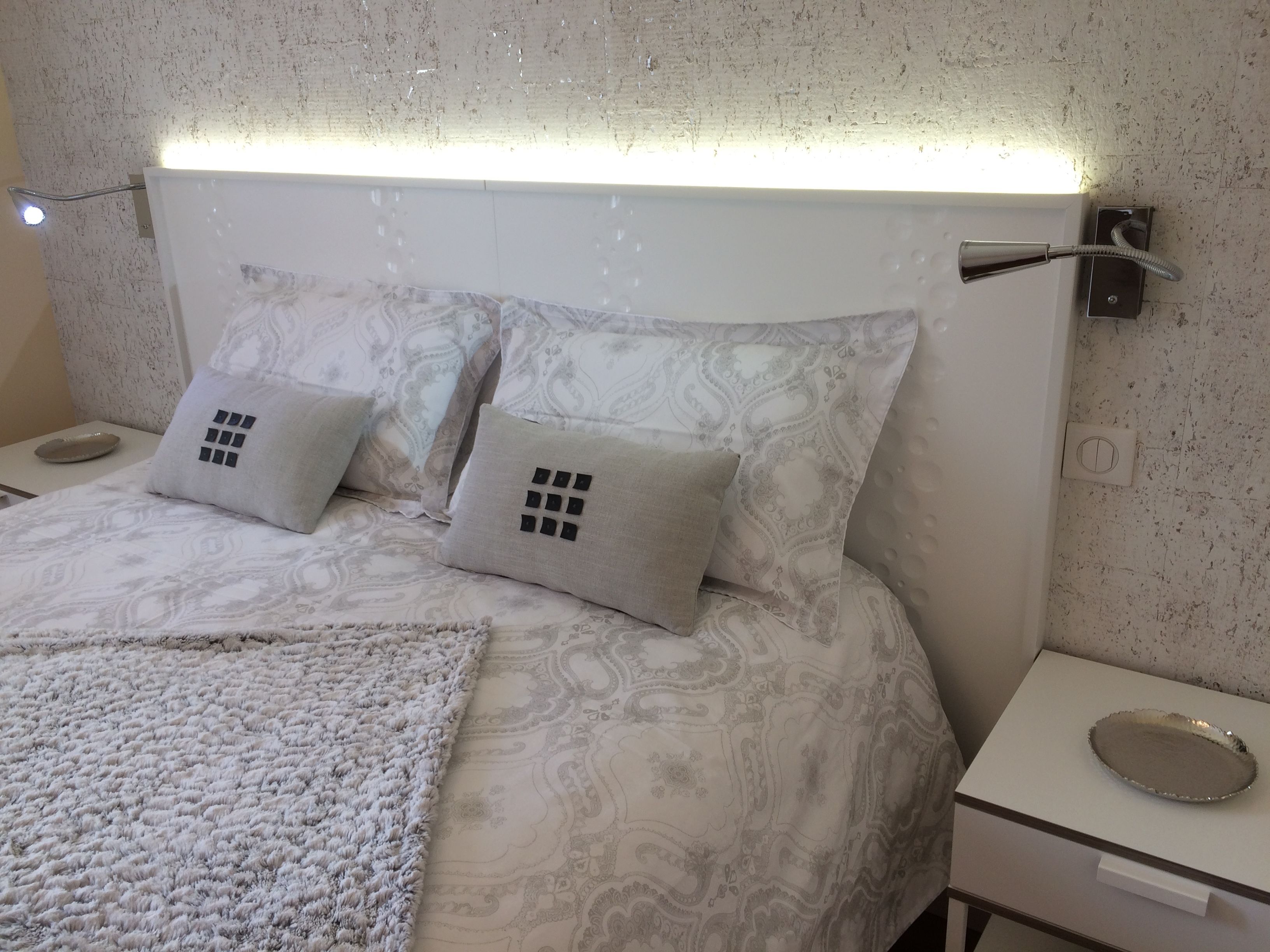 Una cama bien hecha con ropa de cama estampada, dos almohadas con diseño de cuadrícula, una mesita de noche con bandeja y una lámpara de pared.