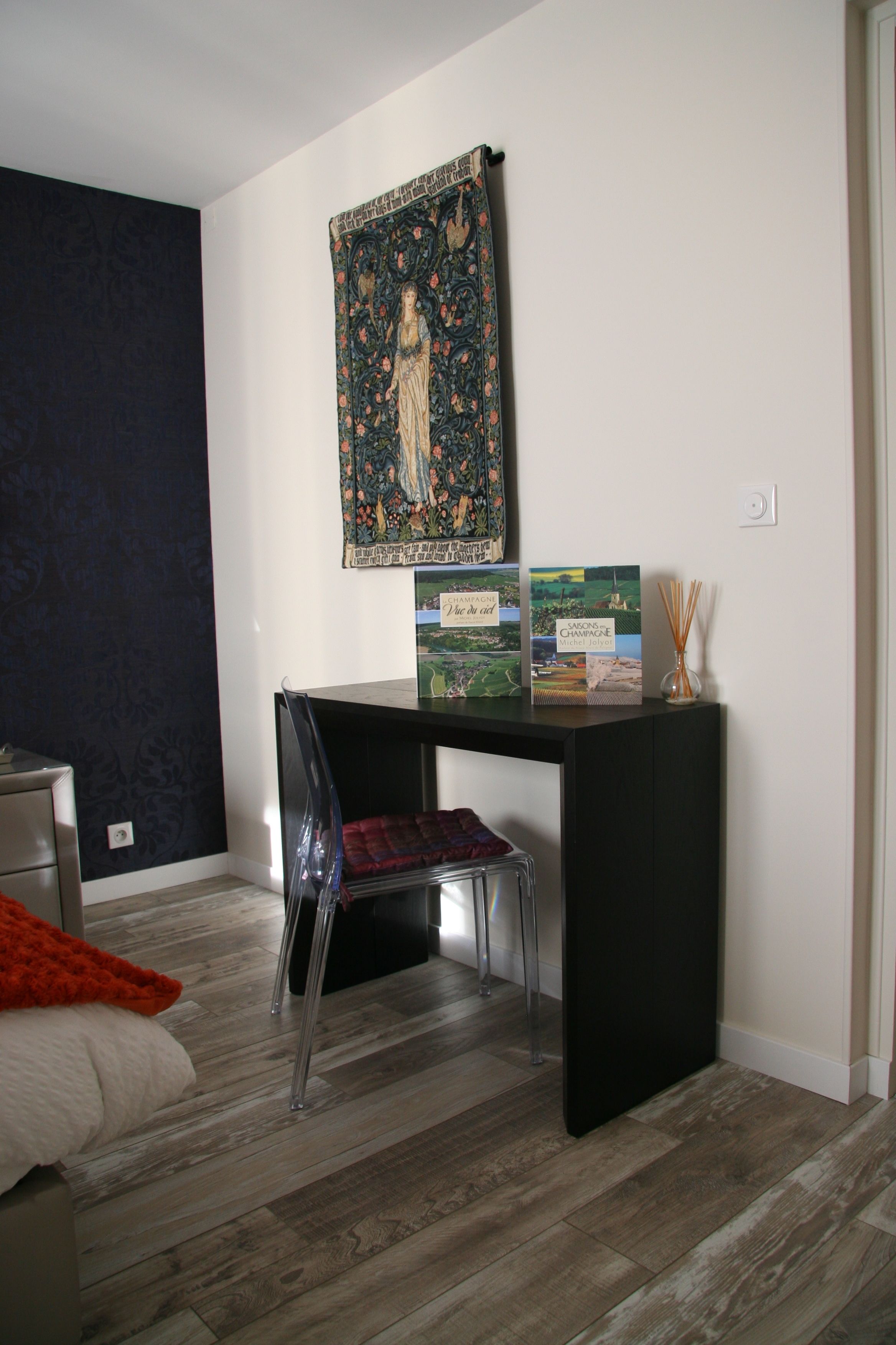A bedroom with a bed, a desk with a chair, a tapestry on the wall, and a small table with books and a candle.