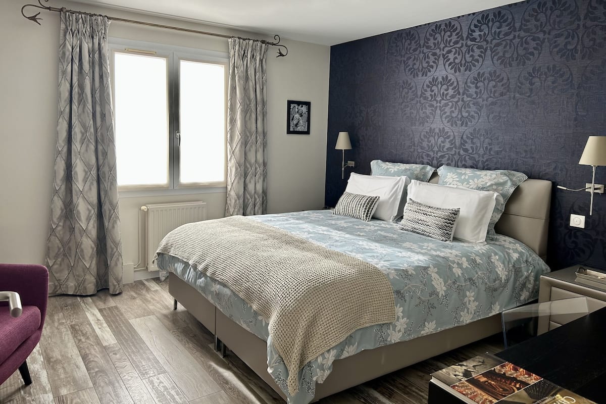 A bedroom with a large bed, two bedside lamps, a window with curtains, a radiator, and a patterned wall.