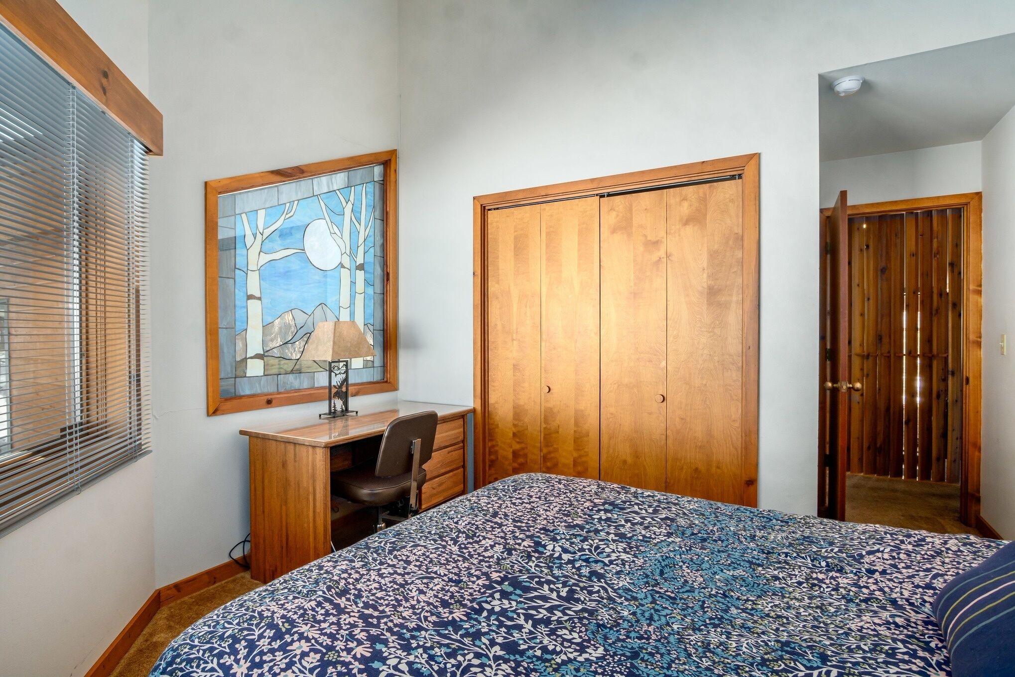 A bedroom with a bed, a desk, a chair, a wardrobe, and a window with blinds.