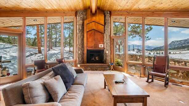 A cozy cabin interior with a stone fireplace, large windows, a sofa, and a dining table. - Stutzhaven in the Mountains - 2 Br Home (Estes Park)