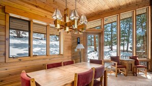 A wooden cabin interior with a dining table, chairs, and large windows offering a view of a snowy landscape. - Stutzhaven in the Mountains - 2 Br Home (Estes Park)