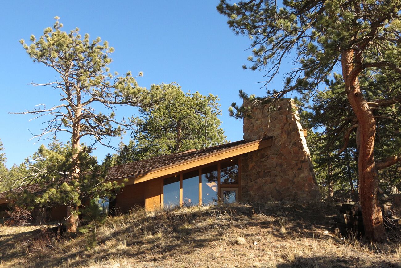 Una casa de piedra con techo inclinado, rodeada de pinos y hierba seca.