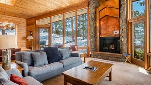 A cozy living room with a stone fireplace, large windows, and a wooden ceiling. - Stutzhaven in the Mountains - 2 Br Home (Estes Park)