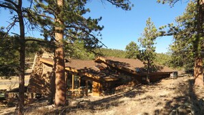 A rustic cabin with a stone foundation, wooden beams, and a sloped roof, surrounded by pine trees and dry grass. - Stutzhaven in the Mountains - 2 Br Home (Estes Park)