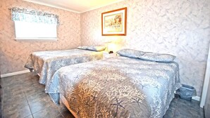 A bedroom with two beds, a window with a curtain, a framed picture on the wall, and a small table with a lamp. - Thunder Island 10A (Ocean City)